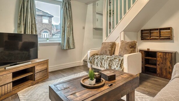 A living room with a TV on a wooden stand, a sofa with a fur throw, a coffee table with a plant, and a staircase with a wooden railing. - Cosy 2BDR Trafford Cottage, Fits 5, EV charger, WIFI, Parking 
 (Partington)