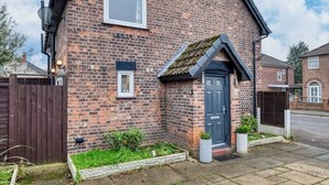 A brick house with a dark blue door, a small window, and a garden area with plants and a white planter. - Cosy 2BDR Trafford Cottage, Fits 5, EV charger, WIFI, Parking 
 (Partington)
