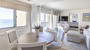 Une pièce avec une table ronde en bois, des chaises blanches, un canapé blanc et un pouf gris. Une grande fenêtre offre une vue sur l’océan.