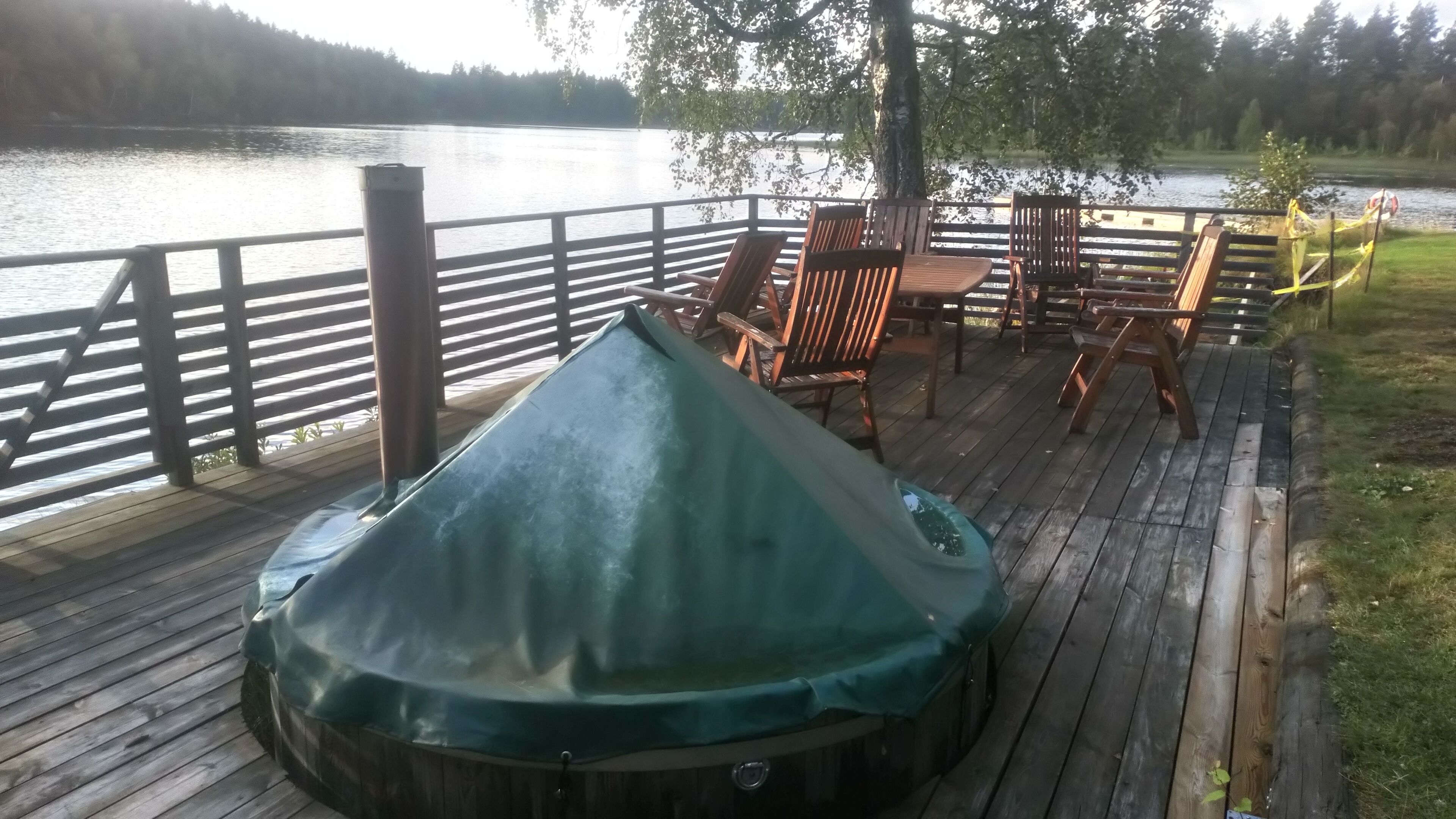 Een houten terras met een overdekte hottub, stoelen en uitzicht op een meer.