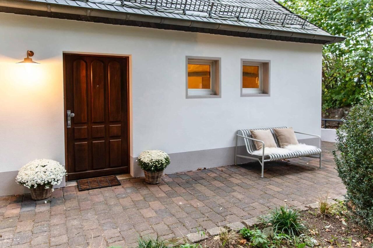 Ein Haus mit einer dunkelholznen Tür, einer weißen Fassade und einem eingepflasterten Eingangsbereich. Neben der weißen Bank mit Kissen steht ein kleiner Blumentopf.