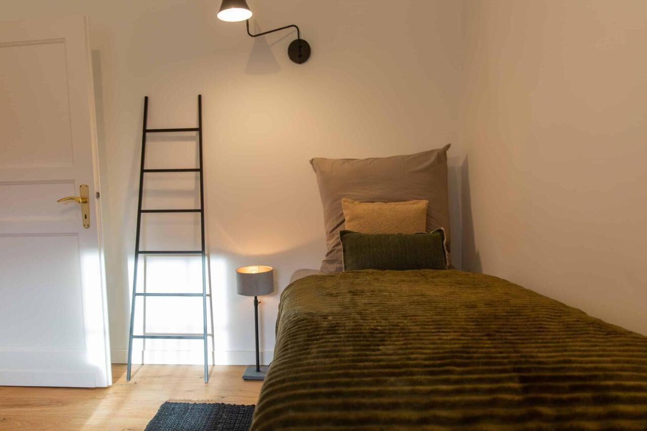 Ein Schlafzimmer mit Bett, Kissen, einer Leiter, einer Stehlampe und einer Tür.