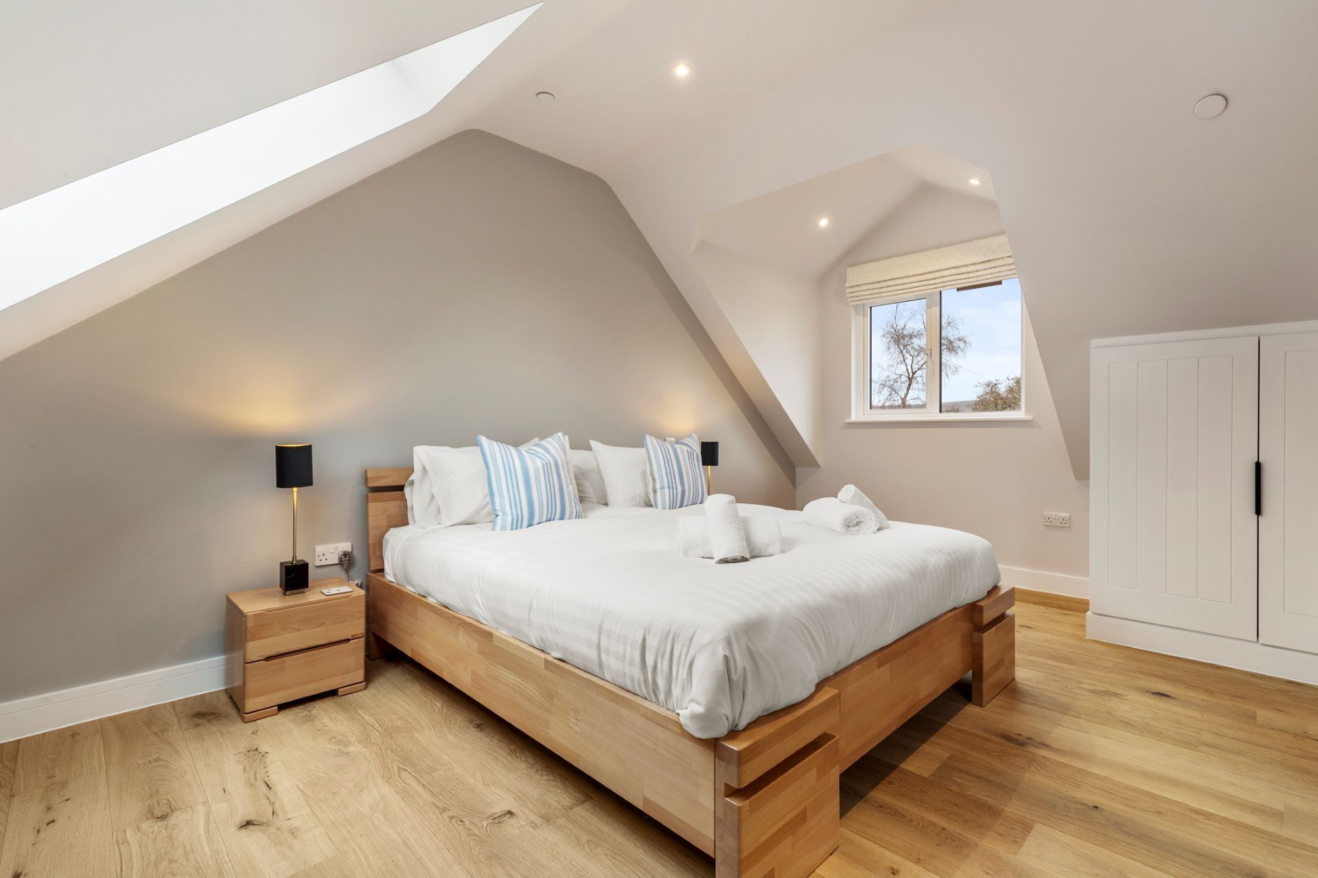 Ein Schlafzimmer mit schrägen Wänden, einem großen Bett, Holz-Nachttischen und einem Fenster mit Jalousien.