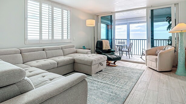 A living room with a sectional sofa, a small table, and a chair. There is a balcony with a view of the sky.