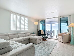 A living room with a sectional sofa, a small table, and a chair. There is a balcony with a view of the sky.