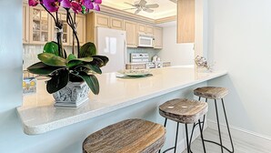 A modern kitchen with white cabinets, a central island with stools, a ceiling fan, and a potted plant with purple flowers.
