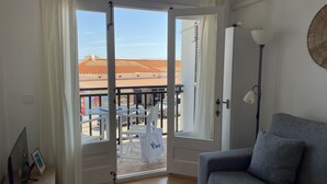 Een kamer met een grijze bank, een witte deur die leidt naar een buitenruimte met tafels en stoelen, een lamp en een wandlamp.