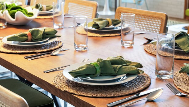 Una mesa de comedor dispuesta con platos, cubiertos y vasos, con individuales tejidos y servilletas verdes.
