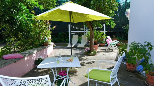 An outdoor patio with white metal chairs and a table, surrounded by potted plants and a yellow umbrella. - I Giardini di Daphne (Meta)