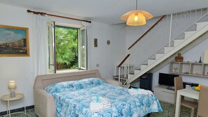 A bedroom with a bed, a desk, a chair, a television, a staircase, and a window with a view of greenery. - I Giardini di Daphne (Meta)