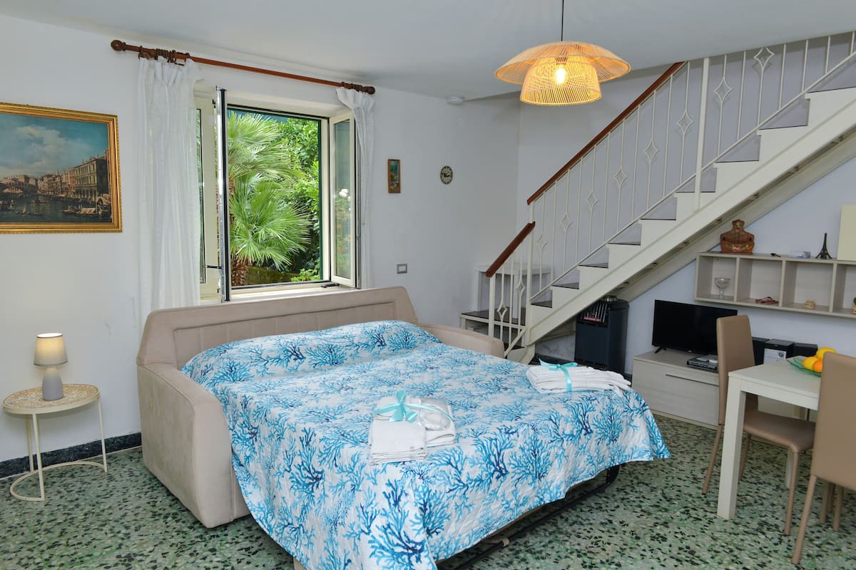A bedroom with a bed, a desk, a chair, a television, a staircase, and a window with a view of greenery.