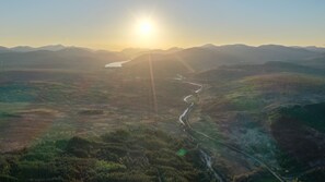 Ein Fluss schlängelt sich durch ein Tal, im Hintergrund Berge und die Sonne, die aufgeht oder untergeht.