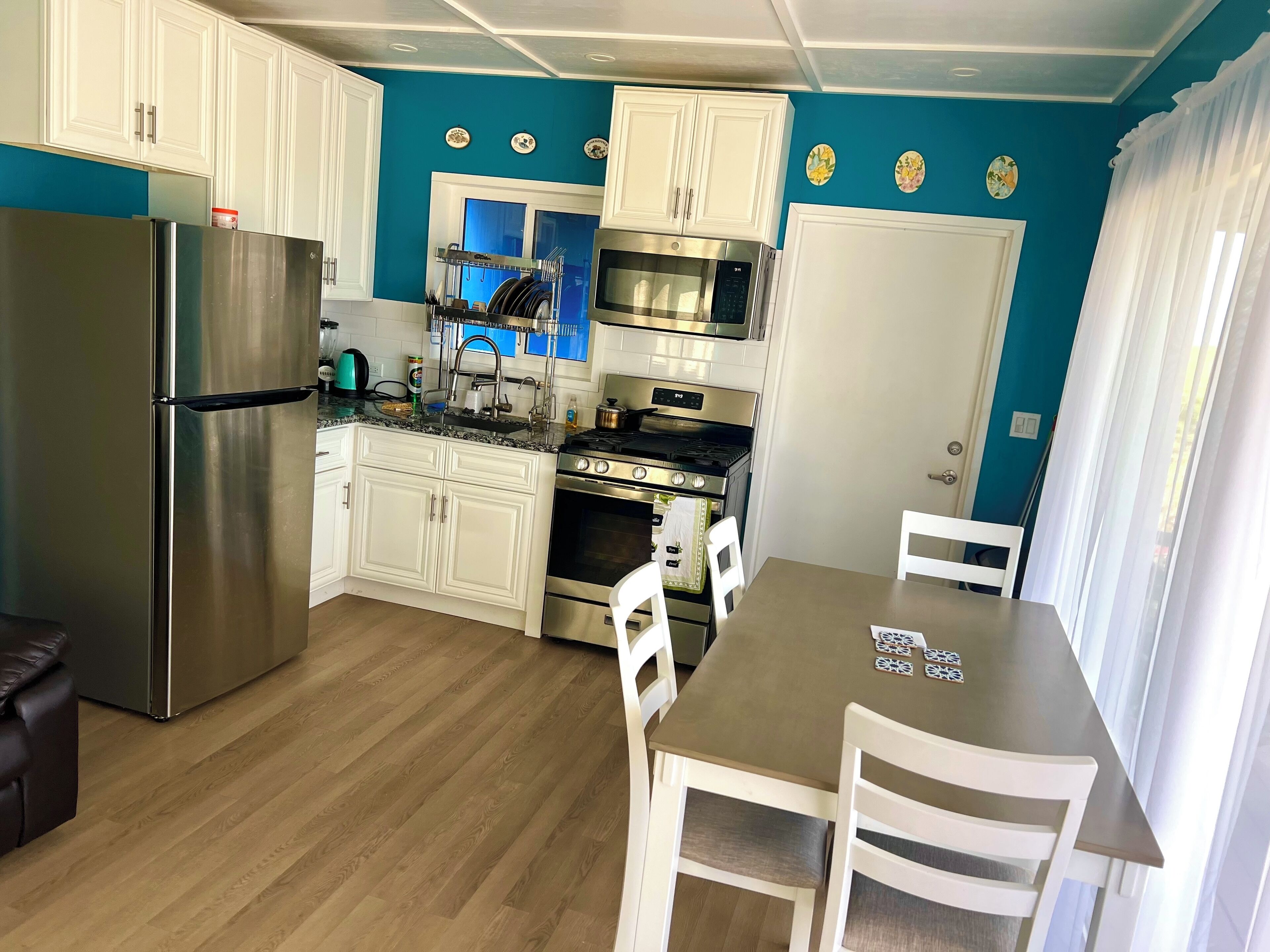 A modern kitchen with white cabinets, a stainless steel refrigerator, a microwave, and an oven. There is a dining table with chairs and a window with curtains.