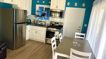 A modern kitchen with white cabinets, a stainless steel refrigerator, a microwave, and an oven. There is a dining table with chairs and a window with curtains.