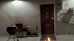 Een vuurkuil in het midden van een betonnen terras, met een barbecue en een opklapbare stoel op de achtergrond.