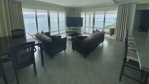 A spacious living room with a large TV, comfortable seating, and a view of the ocean.