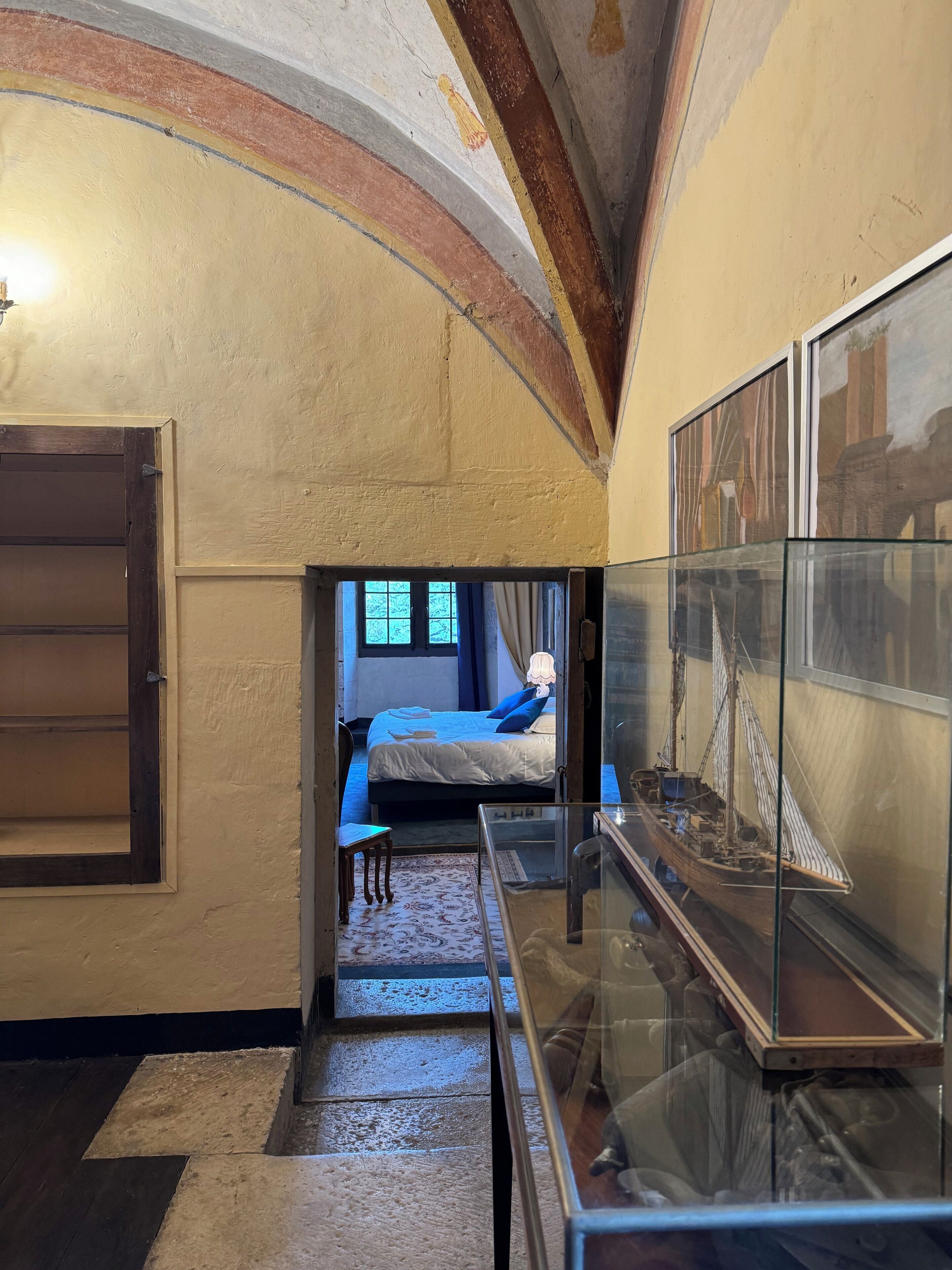 A narrow hallway with a bed in the background, a glass display case with a model ship, and a wooden door on the left.