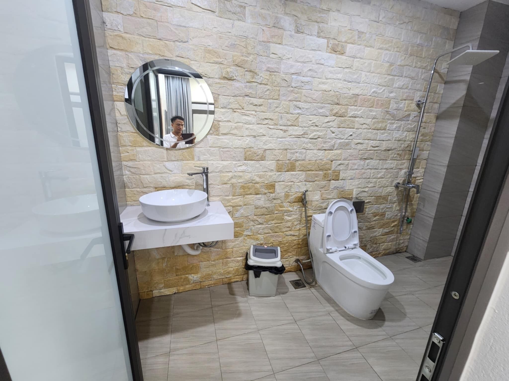 Une salle de bain avec un mur en pierre, un miroir rond, un lavabo, des toilettes et une douche.