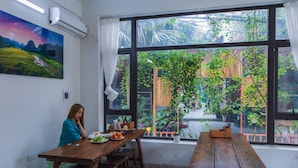 A dining area with wooden tables and benches, a painting on the wall, and a view of a garden through large windows. - Vu's Homestay Tam Coc (Nam Hoa Lu)