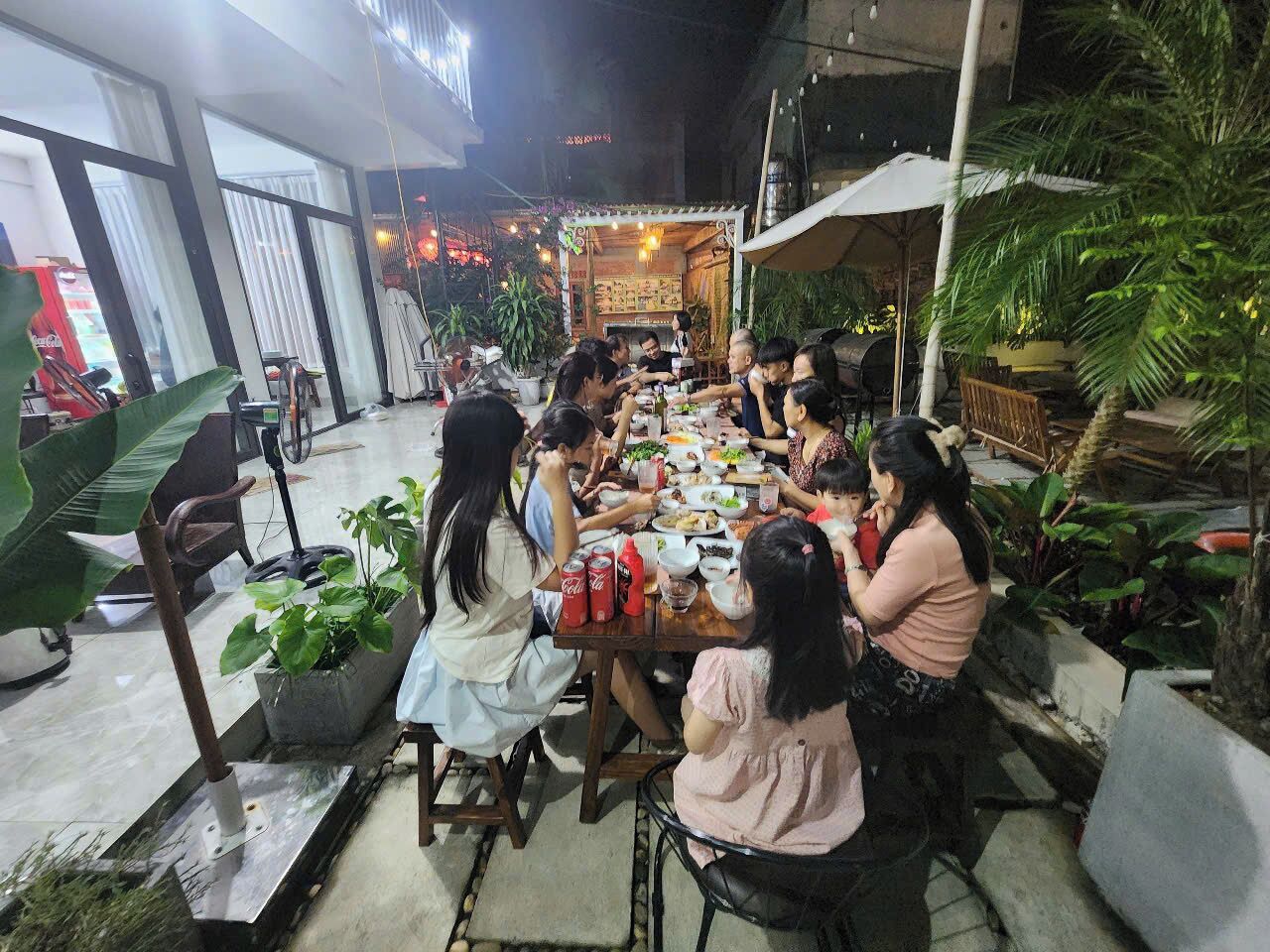 A group of people dining at an outdoor table with various dishes and drinks, surrounded by potted plants and a building in the background.