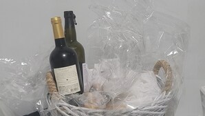 Una cesta de mimbre con una botella de vino y un objeto envuelto, sobre una superficie blanca.