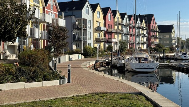 A row of colorful houses along a waterfront with a paved walkway and boats docked nearby.