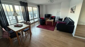 A modern living room with a wooden dining table, two armchairs, a sofa, a TV, and a large window with curtains.