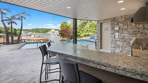 An outdoor kitchen area with a stone wall, a pool, and a view of the surrounding landscape. - Luxury Lakefront Home with Heated Pool and Protected Cove (Sunrise Beach)