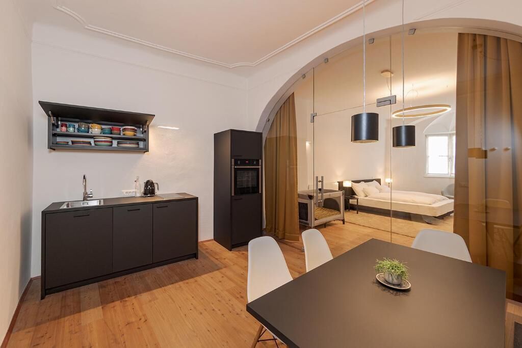 Una cocina moderna con encimera negra, armarios blancos y un estante empotrado con vajilla colorida. Hay una zona de comedor con mesa y sillas, separada por un panel de vidrio de una sala de estar con sofá y lámpara.