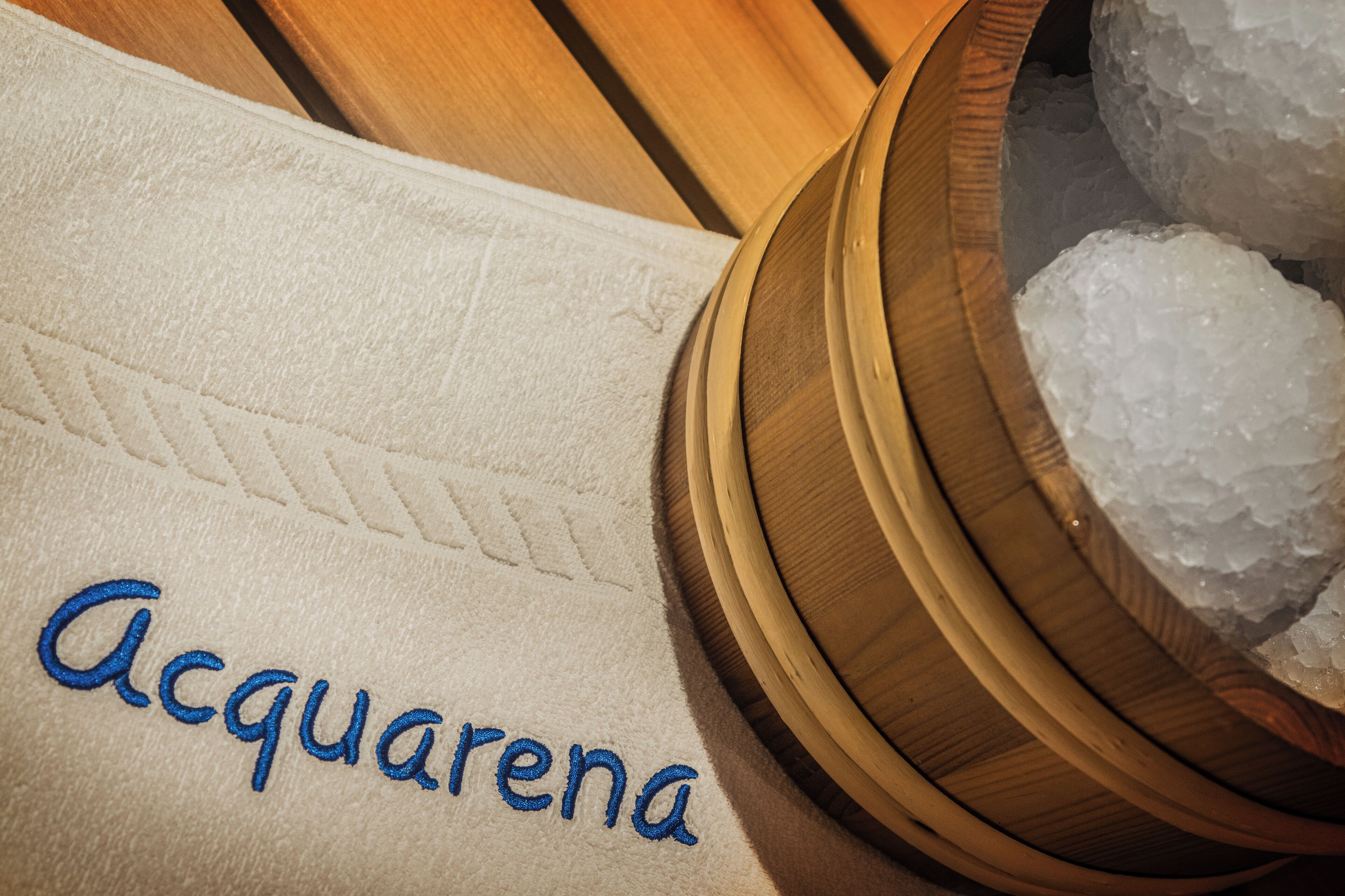A towel with "Acquarena" embroidered in blue, a wooden bowl with ice cubes, and a wooden surface.