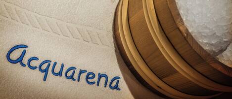 A towel with "Acquarena" embroidered in blue, a wooden bowl with ice cubes, and a wooden surface.