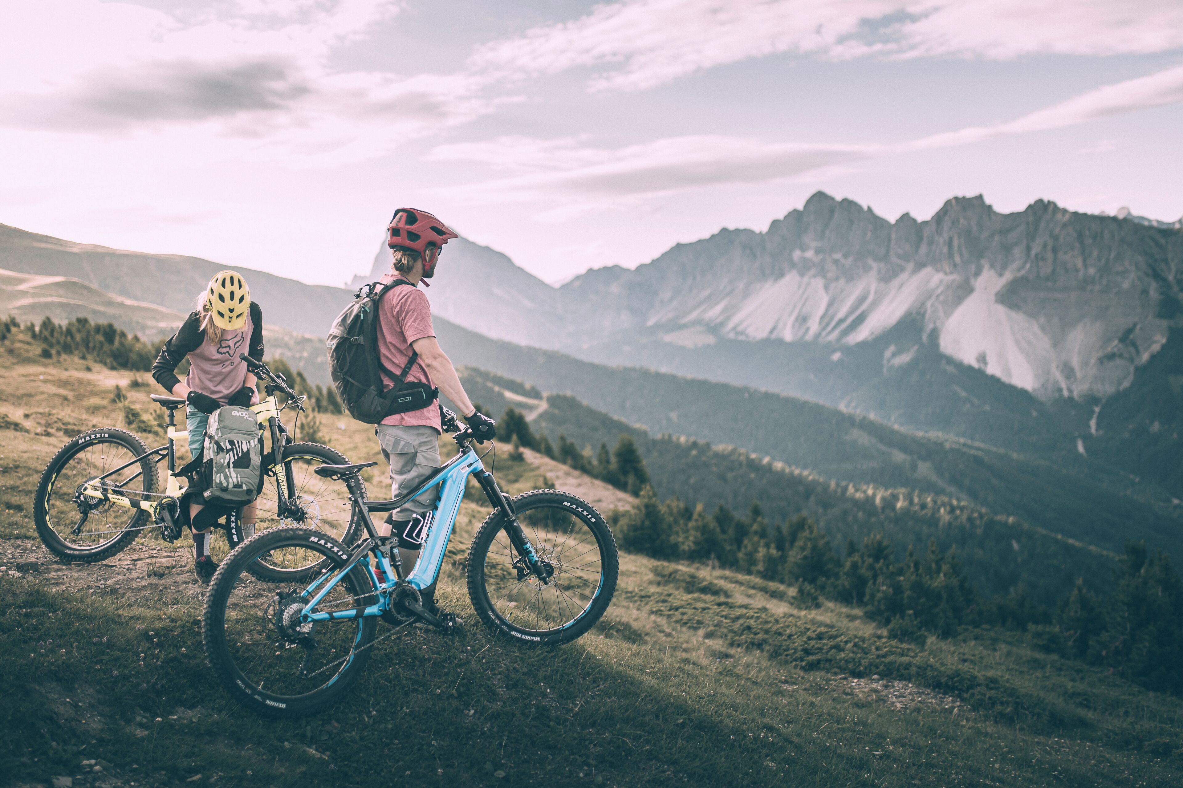 Two people standing on a grassy hill with bicycles, overlooking a mountainous landscape.