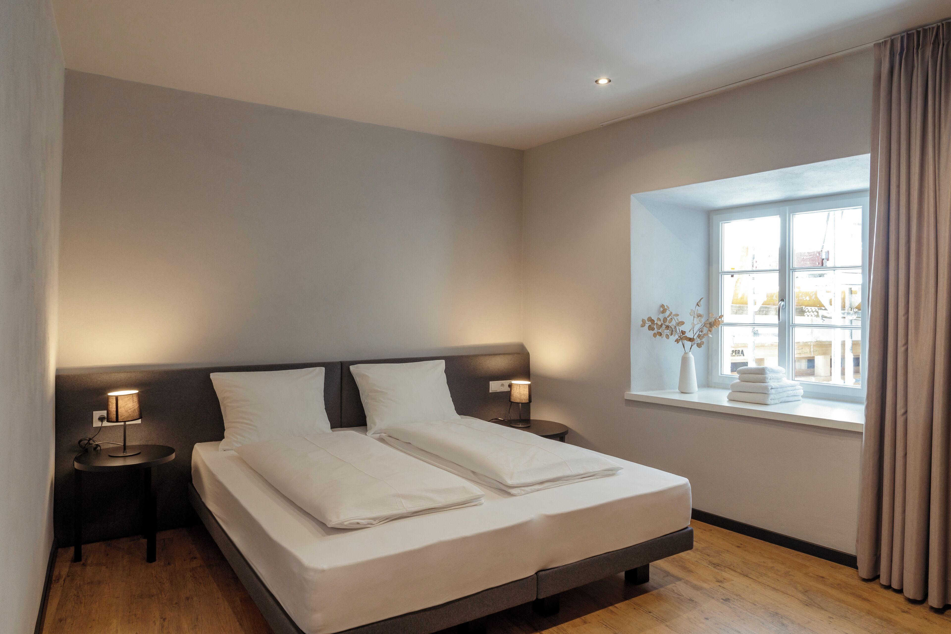 Un lit bien fait, avec du linge de lit blanc, deux tables de chevet avec des lampes, une fenêtre avec des rideaux et une vue sur les bâtiments à l’extérieur.