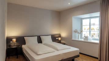 Un lit bien fait, avec du linge de lit blanc, deux tables de chevet avec des lampes, une fenêtre avec des rideaux et une vue sur les bâtiments à l’extérieur.