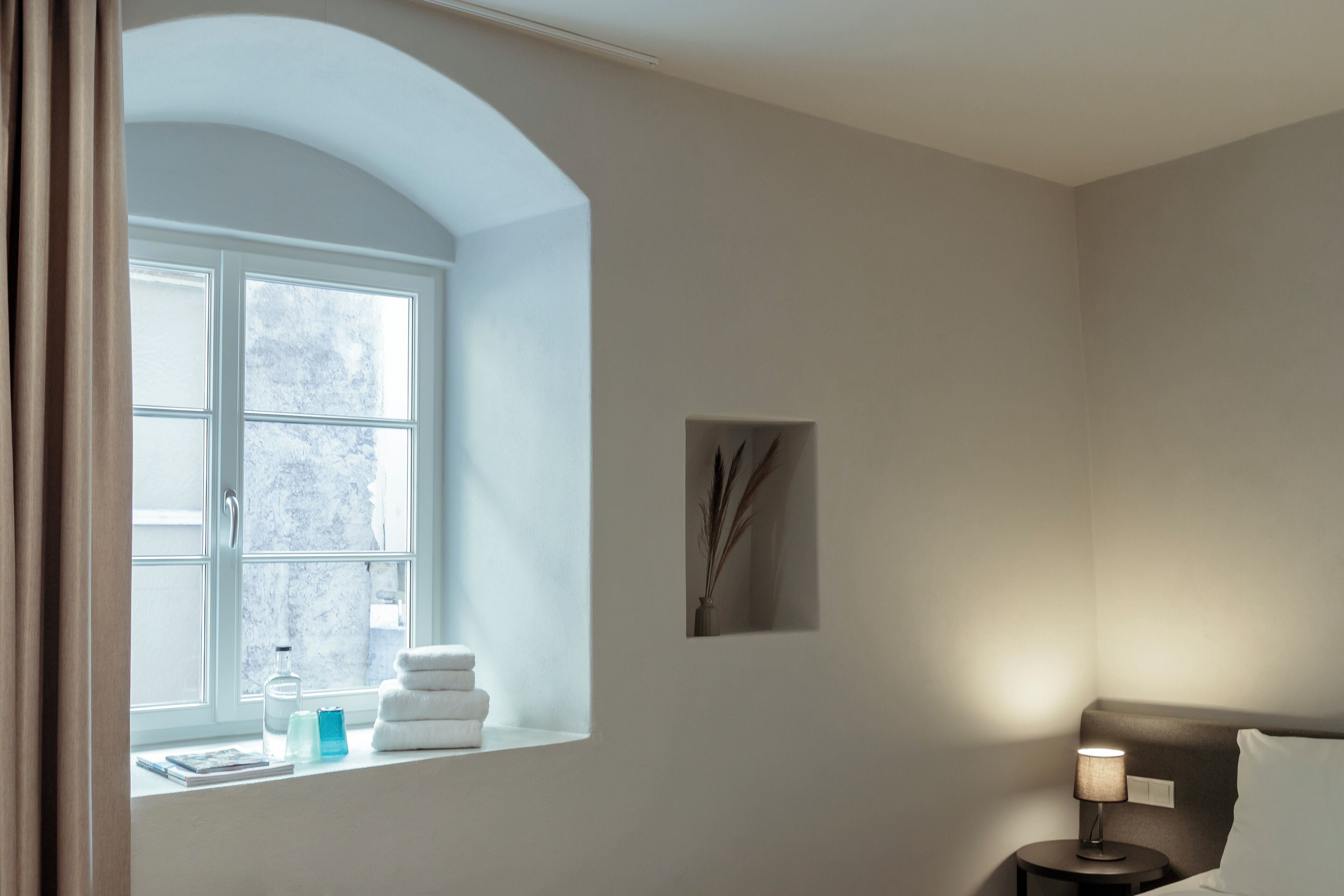 Una habitació d'hotel amb una finestra gran d'arc, una taula de nit amb una làmpada i un quadre penjat a la paret.