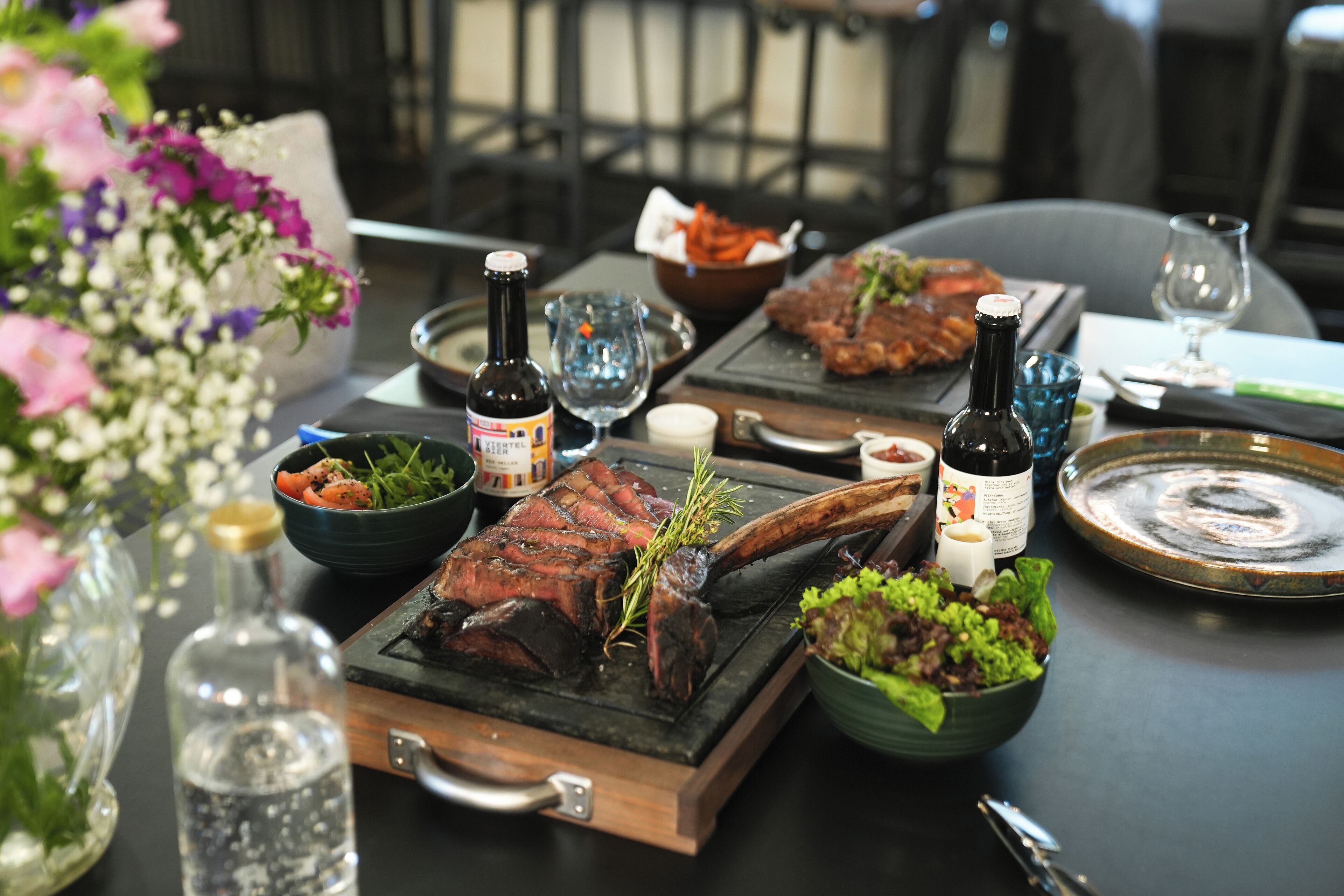 A dining table with a variety of dishes, including a steak, salads, and a bowl of vegetables. There are also several bottles and glasses on the table.