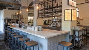 A bar area with a long counter, multiple stools, shelves with glasses and bottles, and a menu board. - Alter Schlachthof (Bressanone)