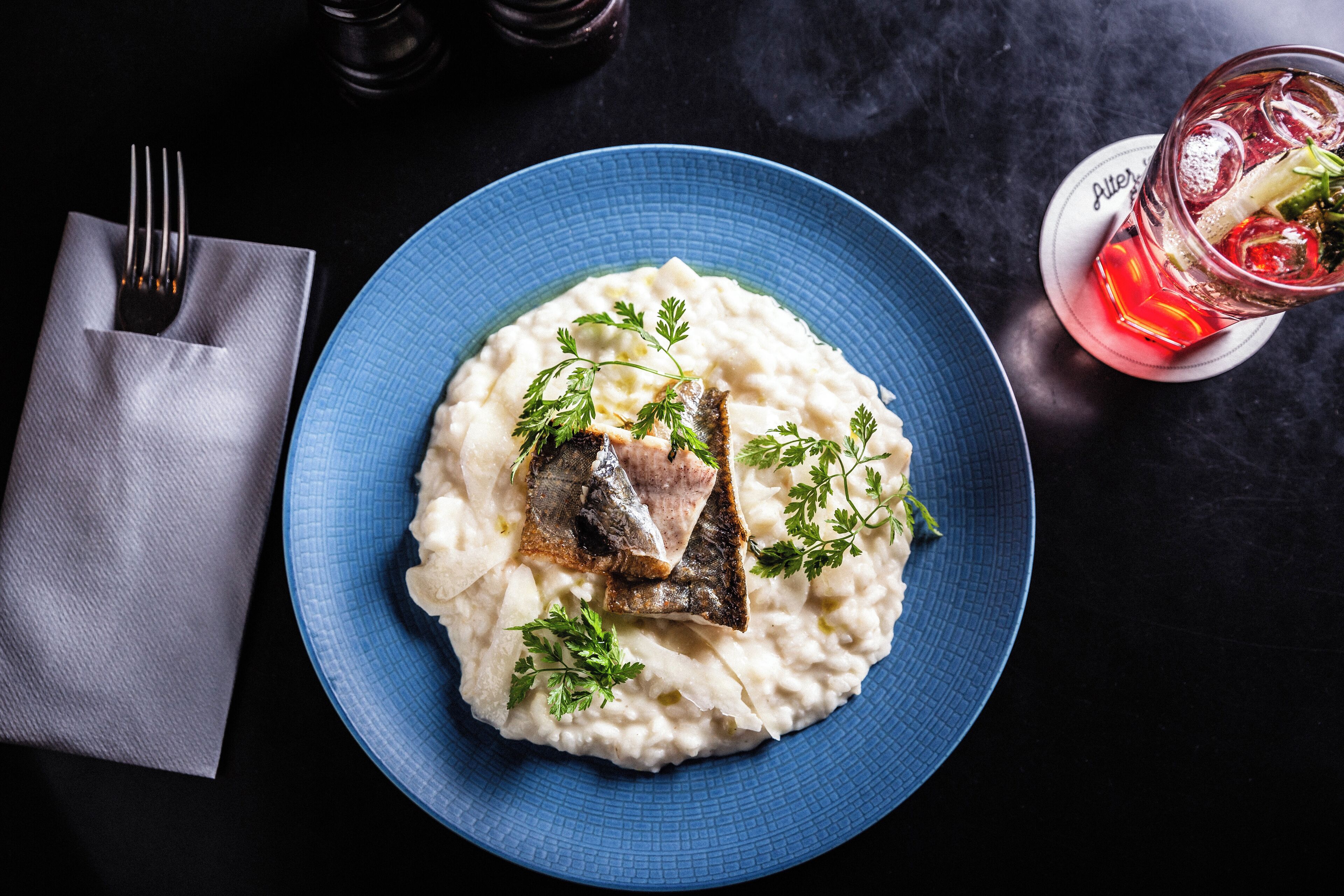 Un plato azul con puré de papas y un trozo de pescado, adornado con perejil, sobre una superficie oscura.
