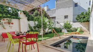 Un patio al aire libre con piscina, sillas rojas y verdes, una mesa con un bol y una mesita con una planta.