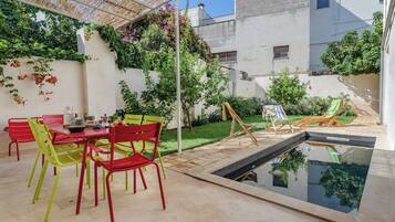 An outdoor patio with a pool, red and green chairs, a table with a bowl, and a small table with a plant.