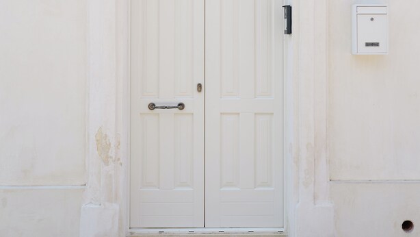 Una puerta doble blanca con un pequeño ventanal encima, en una pared lisa con el número 13 sobre la puerta.