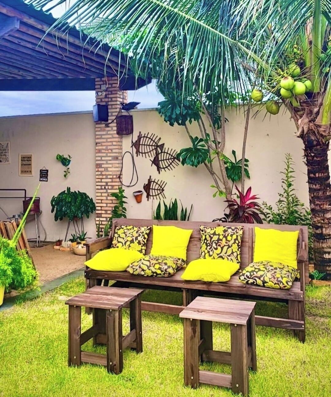 A wooden bench with yellow cushions, a brick wall, and various plants.