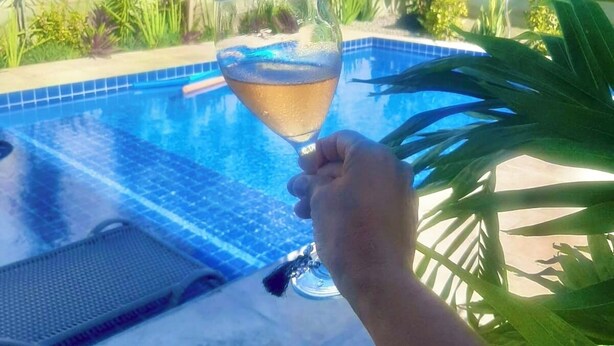 A hand holding a glass of drink over a swimming pool with a view of a garden and a building in the background. - Pousada Areias de Flecheiras (Trairi)