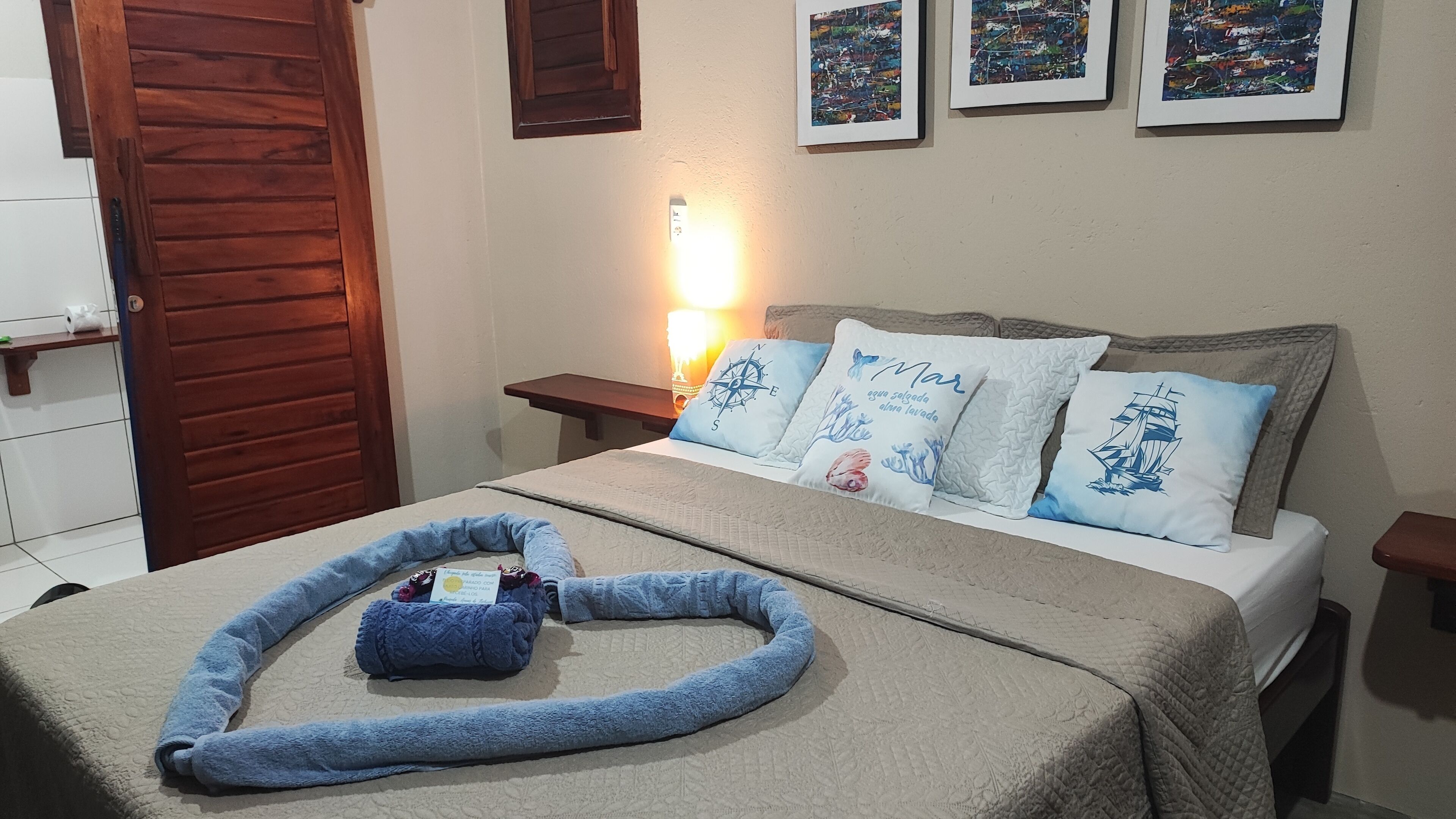 Una cama bien hecha con una manta azul y una toalla azul, una mesita de noche con una lámpara, y tres cuadros en la pared.