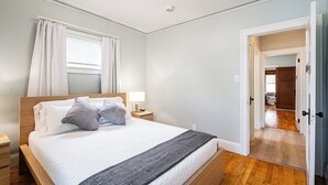Un dormitorio con cama de madera, ropa de cama blanca, una mesita de noche con lámpara y vista a otra habitación por una puerta abierta.