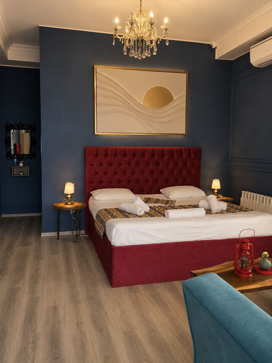 A hotel room with a large bed, a chandelier, a painting, a red upholstered headboard, a bedside table with a lamp, and a red lantern.