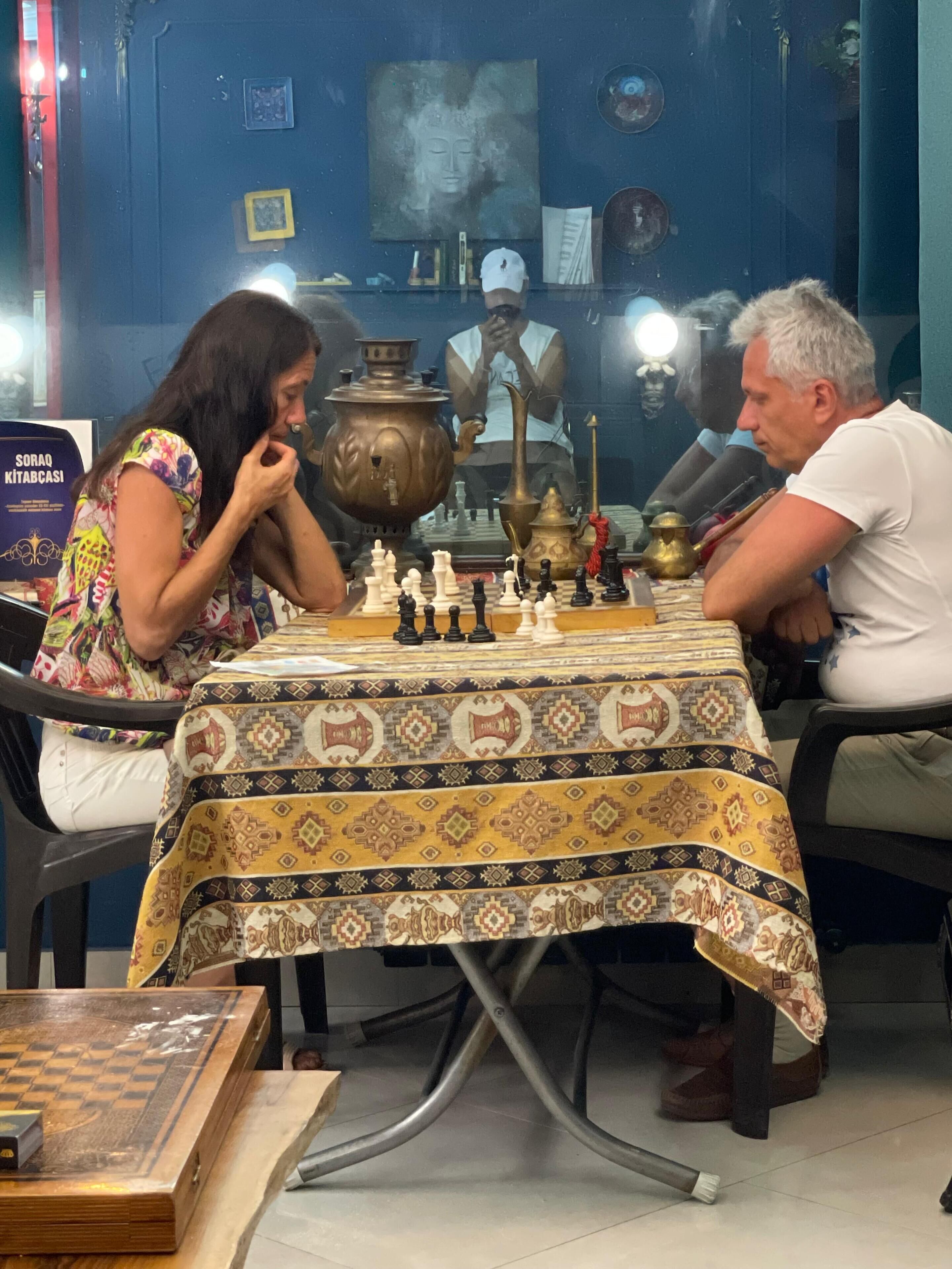 A chessboard set on a table with a patterned tablecloth, surrounded by various decorative items and a statue in the background.