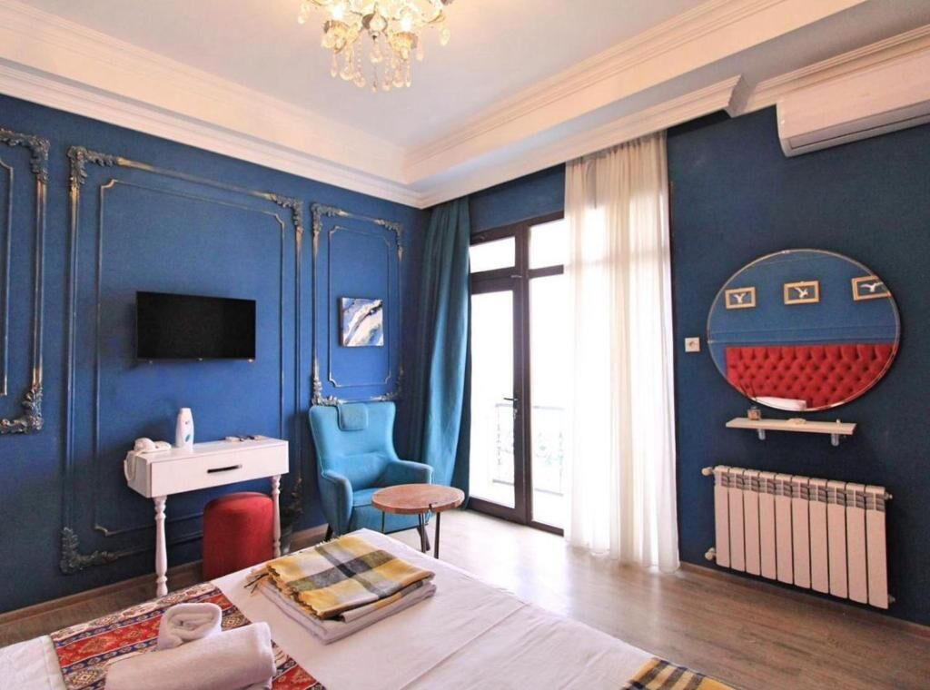 A hotel room with a blue wall, a round mirror, a television, a bed with a red pillow, a chair, a small table, and a radiator.
