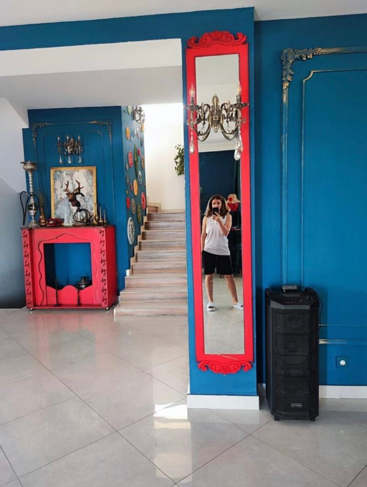 A room with a blue and red color scheme, a large mirror, a fireplace with decorative items, and a staircase leading to another area.