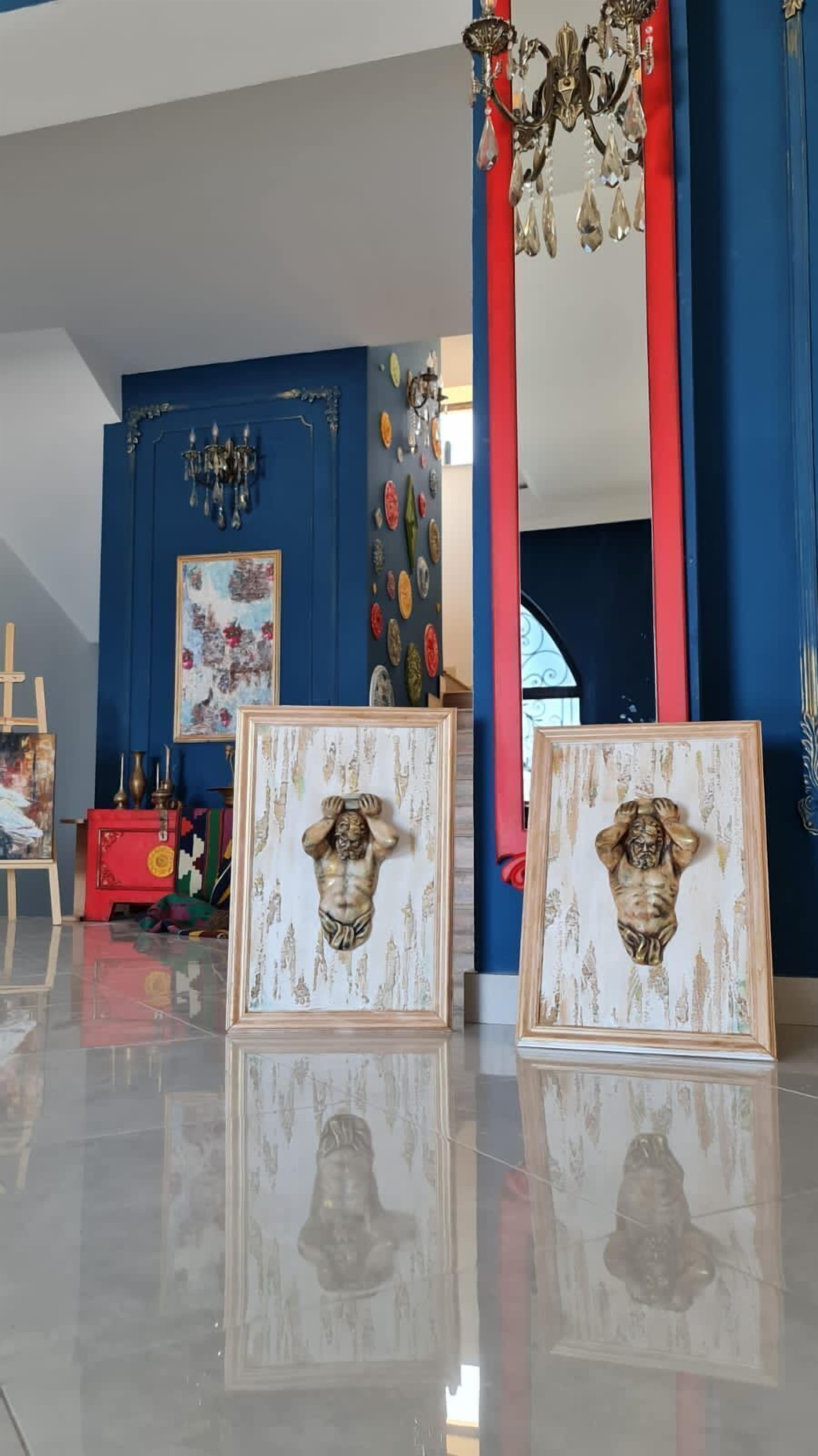 Une pièce avec un mur bleu, un pilier rouge et un lustre. Deux tableaux encadrés sont posés sur le sol.